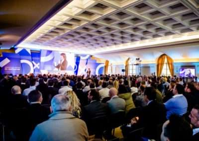 Nell'immagine si possono vedere centinaia di persone assistendo ad un evento. Sullo sfondo un palco per i relatori dove spiccano i colori bianco e una varietà dei blu e celeste. Tuttto allestito da Key Comunicazione.