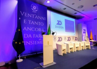 L'immagine rappresenta altro angulo del palco del Congresso e stand dell'Albergatore Day alla Fiera di Roma. E un palco con un sfondo illumaniato che porietta il vido immagine del Sindaco di Roma in cui predominano diverse sfumature del colore blu. Ci sono sedie per partecipanti in colore bianco. E'possibile vedere anche le sagome degli assitenti che guardano verso il palco. Progetto, realizzazione e allestimento per Key Comunicazione.