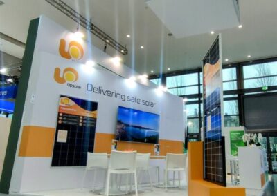 Lo stand Upsolar-HyPower KEY Rimini, realizzato da Key Comunicazione nell' Immagine di uno stand espositivo di Upsolar e HyPower all'interno di un evento. Lo stand è ampio e aperto, con pareti bianche e accenti arancioni. In primo piano, un pannello solare policristallino è esposto. Sullo sfondo, grandi grafiche illustrano paesaggi naturali e promuovono l'energia solare sicura. Ci sono tavoli e sedie bianche per i visitatori e un bancone informativo dove il personale può assistere i partecipanti dell'evento.