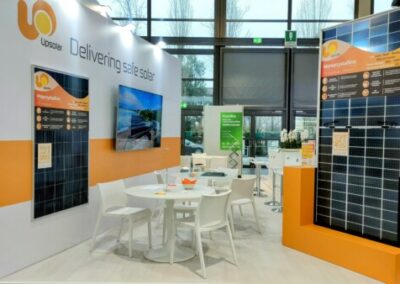 Lo stand Upsolar-HyPower KEY Rimini, realizzato da Key Comunicazione nell' Immagine di uno stand espositivo di Upsolar e HyPower all'interno di un evento. Lo stand è ampio e aperto, con pareti bianche e accenti arancioni. In primo piano, un pannello solare policristallino è esposto. Sullo sfondo, grandi grafiche illustrano paesaggi naturali e promuovono l'energia solare sicura. Ci sono tavoli e sedie bianche per i visitatori e un bancone informativo dove il personale può assistere i partecipanti dell'evento.