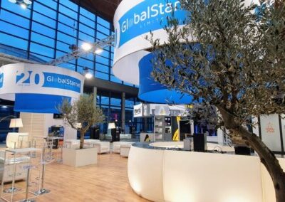 Uno stand elegante con il logo di Globalstarnet, celebrante il 20° anniversario. Al centro, un olivo in un vaso quadrato, illuminato da luci soffuse. Il pavimento è in parquet chiaro e gli arredi includono sedie moderne bianche e tavoli alti con sgabelli.