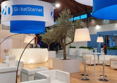 Questa immagine mostra lo stand da una prospettiva più larga, evidenziando il vasto spazio aperto e l'armonia tra i diversi elementi di design come l'illuminazione, i mobili e la vegetazione.