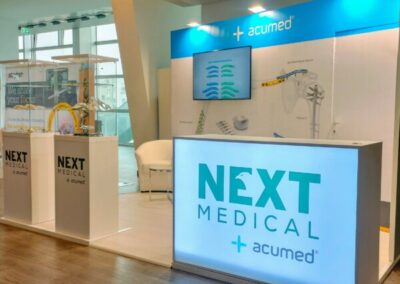 L’immagine mostra uno stand espositivo con il marchio ‘NEXT MEDICAL + acumed’ illuminato su un pannello frontale. Lo stand è composto da pareti bianche e blu con grafiche e testi relativi a prodotti medici ortopedici. Ci sono due vetrine trasparenti che espongono vari strumenti medici e protesi. Sul lato sinistro dello stand, c’è un pannello con il logo di ‘Next Medical’ sopra alcune immagini di spalle e gomiti umani, presumibilmente in relazione alla chirurgia ortopedica. C’è una zona seduta con una poltrona bianca moderna accanto allo stand. L’illuminazione è soffusa ma sufficiente per mettere in evidenza lo stand e i suoi contenuti. Realizzato da Key Comunicazione.