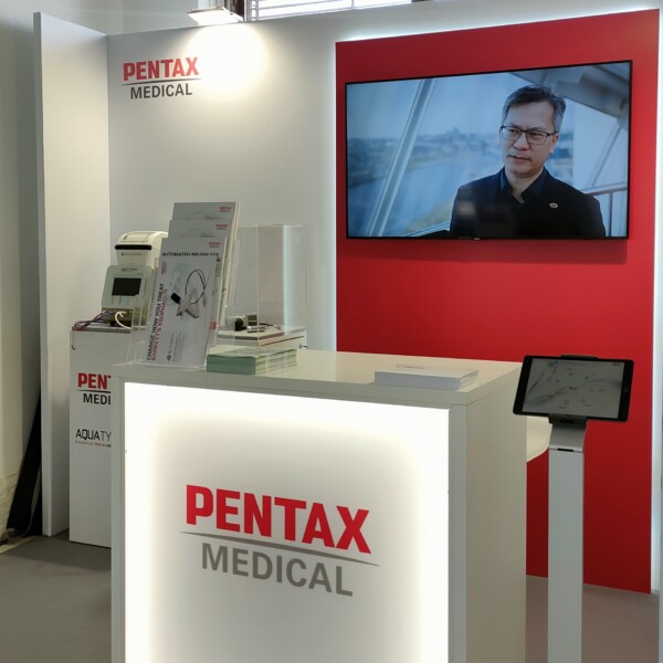 Dettaglio dello stand Stryker al 24° Convegno Nazionale AIIC 2024. In primo piano, un espositore in vetro su un piedistallo bianco con il logo Stryker in nero. Sullo sfondo, letti ospedalieri avanzati e dispositivi medici. Lo stand, illuminato e dai colori bianco e giallo, mette in evidenza le tecnologie sanitarie di Stryker. L'evento si tiene al Congress Center La Nuvola di Roma.