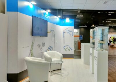 L’immagine si vede lo stand espositivo con il marchio ‘NEXT MEDICAL dove prevale il colore bianco con acceni turchese. illuminato su un pannello frontale. Lo stand è composto da pareti bianche e blu con grafiche e testi relativi a prodotti medici ortopedici. Ci sono due vetrine trasparenti che espongono vari strumenti medici e protesi. Sul lato sinistro dello stand, c’è un pannello con il logo di ‘Next Medical’ sopra alcune immagini di spalle e gomiti umani, presumibilmente in relazione alla chirurgia ortopedica. C’è una zona seduta con una poltrona bianca moderna accanto allo stand. L’illuminazione è soffusa ma sufficiente per mettere in evidenza lo stand e i suoi contenuti. Realizzato da Key Comunicazione.