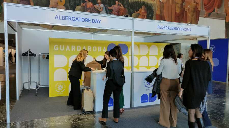 In prospettiva il palco realizzato da Key Comunicazione per conto di Federalberghi Roma in ocassione dell'Albergatore Day 2025. Spica il colore bianto tra diverse tonalità di blue sull0 sfondo del palco dove viene proiettato un video. La platea piena degli assitengi all'evento.