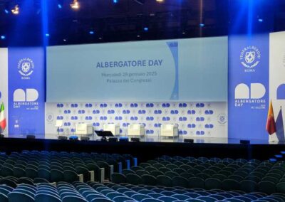 Inaugurazione dell'Albergatore Day 2025 al Palazzo dei Congressi di Roma, con il palco ufficiale allestito da Key Comunicazione, caratterizzato da un design elegante e innovativo, pannelli personalizzati e un’illuminazione scenografica., Lo sfondo dell'scenario ha un maxi schermo su una parete bianca in contrasco con diverse tonalita' di blu come i colori ufficiali dell'evento.