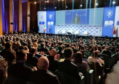 Tante persone assistono alla presentazione del sindaco di Roma Walter Gualtieri sul palco dell'Albergatore Day 2025 al palazzo dei Congressi di Roma. Uno sfondo bianco e blu fa di cornice al maxischermo che progetta l'immagine amplificata del Sindaco di Roma. Il palco per le esposizioni dei relatori dell'Albergatore Day 2025 di Roma. Le sedia della plates in colore verde/blue secondo l'uminazione ed lo sfondo del placo in bianco dove spiccano i loghi della Federalerghi di Roma in colore blue.