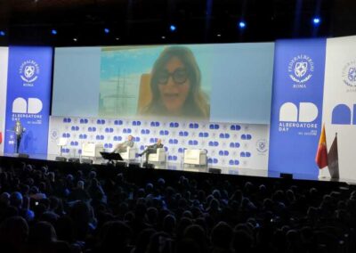 Il maxischermo progetta l'immagine video della Sen. Daniela Santachè dietro il palco con delle persone con uno sfondo bianco e blo conn il logo dell'evento Albergatore Day. Una platea gremita di persone assiste alla presentazione. Allestimento per Key Comunicazione.
