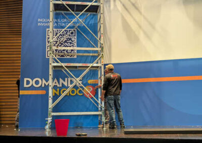 Backstage dell'evento 'Domande in Gioco' di IGT, allestito da Key Comunicazione. Tecnico al lavoro su impalcatura, pannello blu con logo IGT e claim 'Domande in Gioco', dettagli dell'installazione professionale al Palazzo dei Congressi di Roma." Descrizione dettagliata: L'immagine mostra un momento dell'allestimento tecnico, con un operatore in piedi su un'impalcatura metallica. Il pannello blu riporta il logo IGT, un codice QR e il claim "Domande in Gioco". La struttura metallica attraversa verticalmente il pannello, mentre un secchio rosso è posizionato sul palco. L'illuminazione e l'accuratezza dell'installazione rivelano la precisione e la professionalità tipiche di Key Comunicazione nell'organizzazione di eventi aziendali. I dettagli tecnici e l'attenzione alla scenografia sono evidenti in ogni aspetto dell'allestimento.