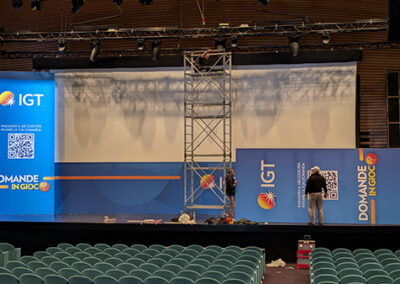 Fase di allestimento dell'evento 'Domande in Gioco' di IGT, curato da Key Comunicazione. Palco con maxischermo blu, struttura in alluminio, tecnico al lavoro e sedie verdi in primo piano. Preparazione professionale dell'evento al Palazzo dei Congressi di Roma." Descrizione dettagliata: L'immagine cattura un momento cruciale dell'preparazione dell'evento. Il palco è in fase di allestimento, con una struttura in tralicci metallici che supporta lo schermo principale. Il design dello schermo riprende l'identità visiva di IGT, con colori blu e arancione, e include il logo dell'azienda e un codice QR. Un tecnico è di spalle, intento a sistemare gli ultimi dettagli. Le sedie verdi in platea sono ancora vuote, in attesa dei partecipanti. L'illuminazione e l'attrezzatura suggeriscono la cura meticolosa di Key Comunicazione nell'organizzazione di eventi aziendali di alto livello.