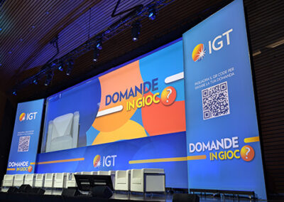 Evento IGT Domande in Gioco al Palazzo dei Congressi di Roma: palco con maxi schermi LED che mostrano il logo IGT e la grafica ufficiale dell'evento. Sedie bianche disposte per i relatori, luci di scena e un QR code per interagire con le domande del pubblico.