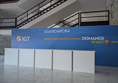 "Guardaroba dell'evento 'Domande in Gioco' di IGT, allestito da Key Comunicazione. Bancone bianco con pannello blu IGT, scritta 'Guardaroba' e logo dell'evento, posizionato in un elegante ingresso con scale e pavimento in marmo." Descrizione dettagliata: L'immagine mostra il punto guardaroba dell'evento, perfettamente in linea con l'identità visiva di IGT. Un bancone bianco pulito e minimale è posizionato sotto un grande pannello blu che riporta il logo IGT, la scritta "Guardaroba" e il claim "Domande in Gioco". Sullo sfondo si vedono scale e colonne in marmo, che conferiscono all'ambiente un'atmosfera elegante e professionale. L'allestimento riflette la cura e l'attenzione ai dettagli che contraddistinguono Key Comunicazione nell'organizzazione di eventi aziendali di alto livello.