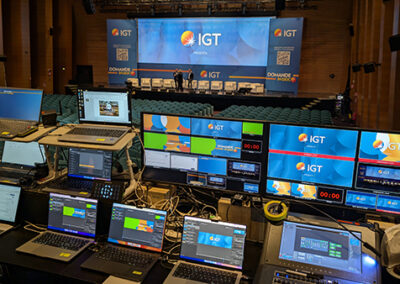 Regia tecnica dell'evento 'Domande in Gioco' di IGT, curata da Key Comunicazione. Numerosi monitor e laptop configurati per la produzione multimediale, con schermi che mostrano il logo IGT e interfacce di controllo. Postazione di regia tecnica professionale allestita per la gestione completa dell'evento al Palazzo dei Congressi di Roma." Descrizione dettagliata: L'immagine mostra la complessa postazione di controllo tecnico dell'evento. Un insieme di laptop e monitor professionali sono disposti su un tavolo nero, con schermi che mostrano diverse interfacce grafiche, loghi IGT e contenuti multimediali. I monitor hanno una predominanza di colori blu e arancione, coerenti con l'identità visiva dell'evento. La disposizione accurata delle attrezzature e la molteplicità di schermi suggeriscono un'organizzazione tecnica accurata e professionale, tipica di Key Comunicazione nella gestione di eventi complessi.