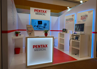 Allestimento Stand Pentax Medical al Congresso FISMAD curato da Key Comunicazione. Inquadratura laterale dell’area espositiva con due schermi attivi che mostrano dispositivi endoscopici. Il bancone reception centrale, retroilluminato, riporta il logo Pentax Medical. L’arredamento minimalista e l’illuminazione LED creano uno spazio accogliente e professionale. Presenti anche elementi naturali come piantine verdi, per rendere l’ambiente più umano e visivamente equilibrato. Lo stand comunica con chiarezza l’orientamento all’innovazione del brand.