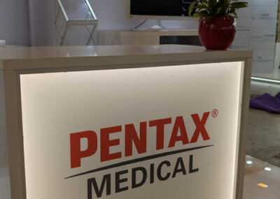 Desk frontale dello stand Pentax Medical all’AIIC firmato Key Comunicazione, con pianta decorativa e monitor di presentazione in sfondo.