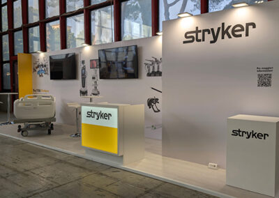 Stand Stryker AIIC Napoli 2025. Stand medicale progettato da Key Comunicazione per Stryker, con banco reception retroilluminato giallo, grafiche tecniche e monitor integrati su parete bianca.