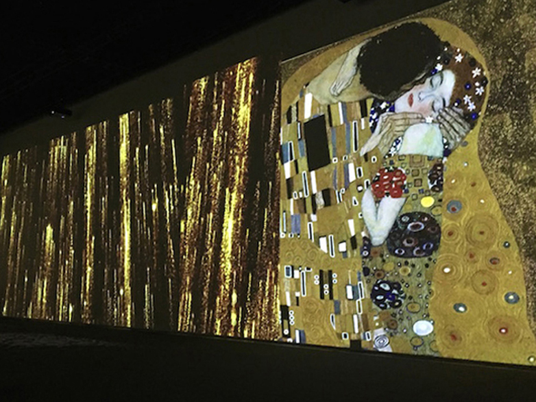 Una visuale dell'allestimento proegettato per la Mostra Interattiva "Klimt Experience" - In quest'immagine, la proiezioni di formati giganti dell'opera 'Il Baccio' di Klimt con sfondi giallo - oro raffigurando una uomo bacciando passionalmente ad una donna. Progette e allestimento a cura di Key Comunicazione.