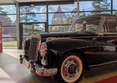 Automobile Mercedes-Benz d'epoca esposta accanto a un pannello informativo, con la Cupola di San Pietro sullo sfondo.